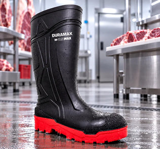 Bota de hule industrial negra con suela roja antiderrapante sobre piso húmedo en una planta procesadora de alimentos.