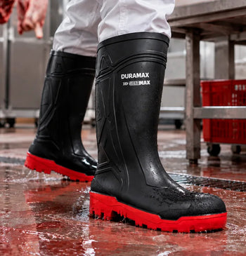 Bota impermeable de hule color negro con suela roja antiderrapante sobre piso mojado en planta de alimentos.