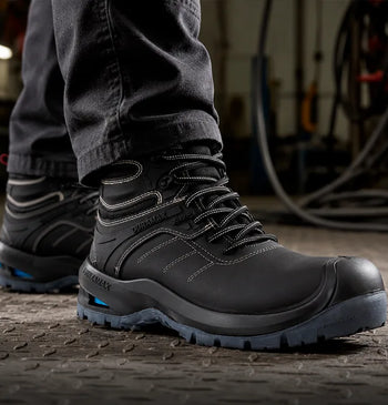 Primer plano de botas industriales negras de corte ergonómico sobre suelo metálico estriado en un taller especializado.